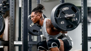 An athlete in a sleeveless gray shirt, black shorts, and kneepads performs squats in the gym with a heavy barbell over their shoulders.