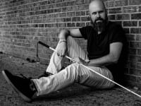 'The Blind Life' host Sam Seavey shown sitting up against a brick wall holding a guide cane.