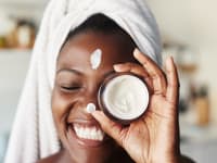 A woman wearing a towel on her head and smiling as she holds up a jar of cream in front of her eye. She has a strip of moisturizer on her forehead and on her nose.