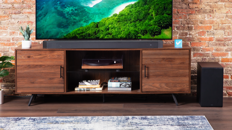 The Samsung HW-Q990C soundbar in front of a TV on a home theater credenza with its sub on the floor to the side.