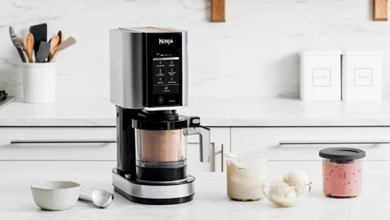 A ninja creami machine sits on a counter with ingredients bowls and an ice cream scoop