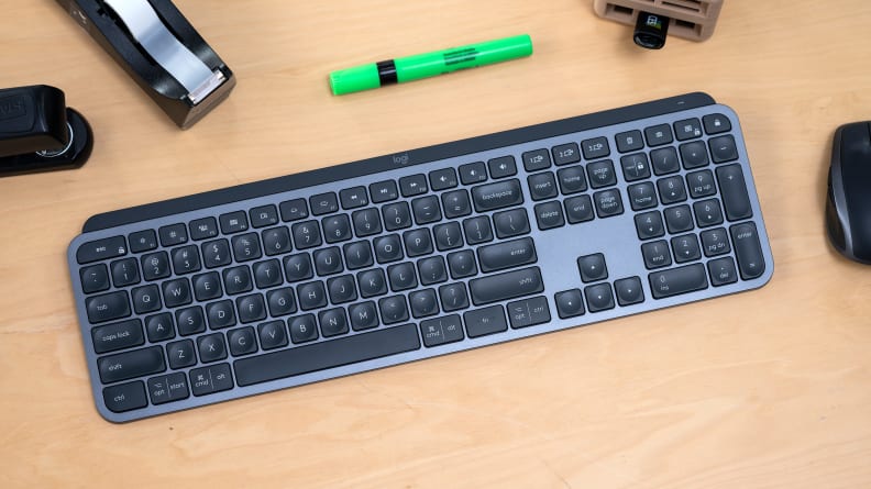 A wireless keyboard rests on a wooden desktop. A stapler, highlighter, tape dispenser, and wireless mouse are all close by.