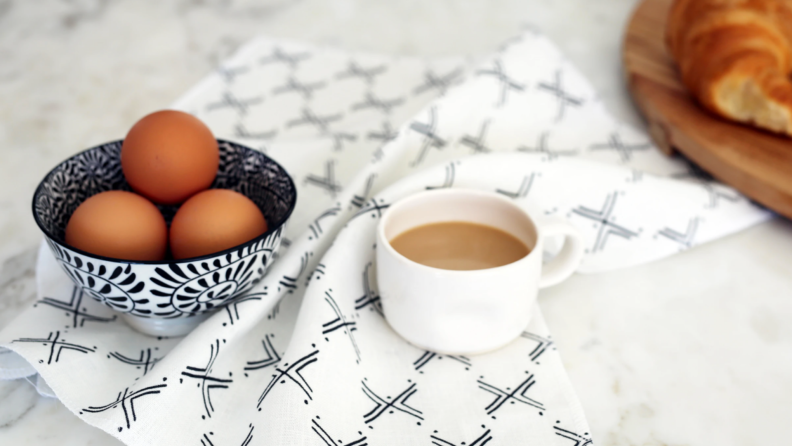 White and black patterned kitchen cloth next to white coffee mug, bowl of eggs, and a cutting board with bread on it