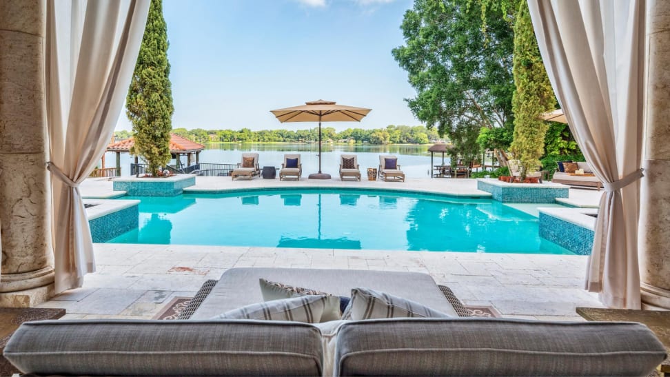 Outdoor living area by a lake facing a large pool with an outdoor couch, outdoor curtains, lounge chairs, and an umbrella