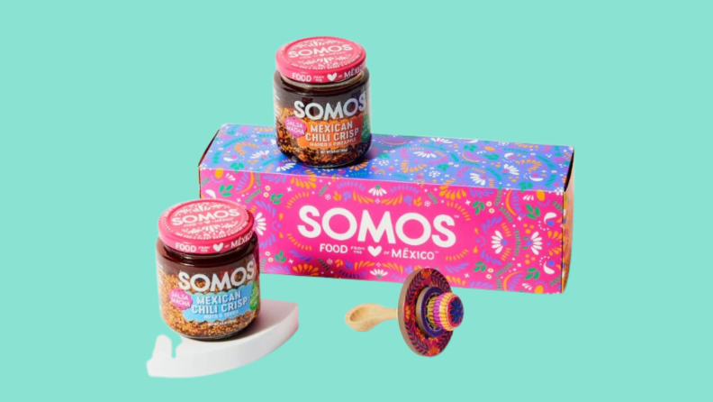 Two jars of Mexican chili crisp from Somos arranged on and near a Somos package and a spoon with a sombrero handle.