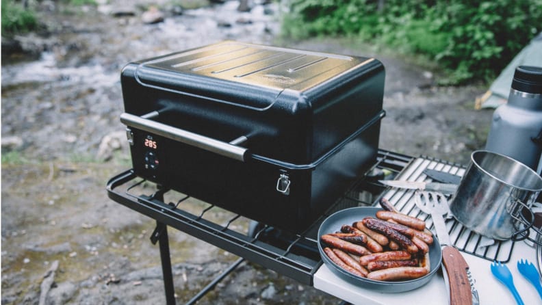 A Traeger portable grill sits on a stand at a campsite.