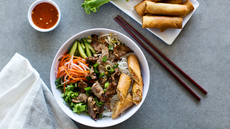A bowl of Vietnamese lemongrass beef noodle salad is in the center of the image. A plate of spring rolls and dipping sauce are next to it.