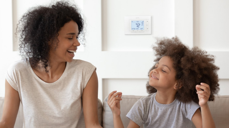 A mom and child sit on a couch with a Sensi smart thermostat hanging on the wall behind them.