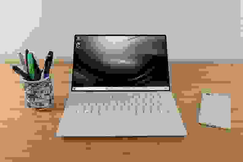 An open laptop on top of a desk with a cup of pens and a phone on either side.