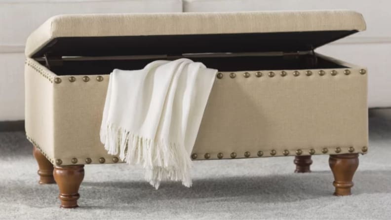 An Oakford upholstered storage bench is pictured with the top lifted up, showing a white throw blanket being put inside (along with whatever else you need to store).