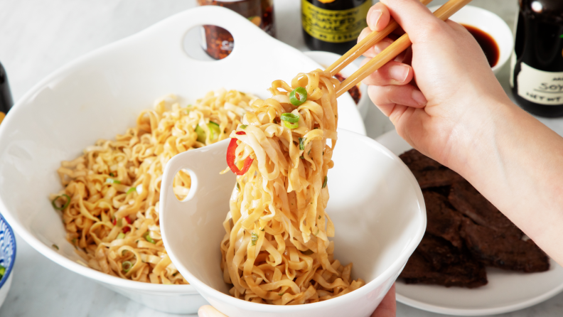 Person using chopsticks to lift cooked noodles inside of white bowl.