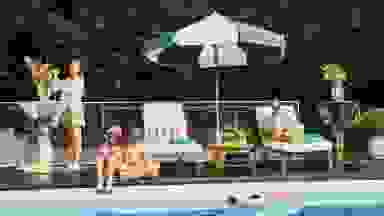 A young family relaxes by the pool under an umbrella while the Aiper Scuba X1 Pro Max cleans the pool nearby