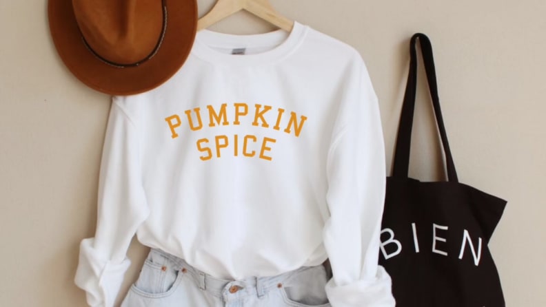 A white sweater that says 'Pumpkin Spice' next to a hat, tote bag, and jeans