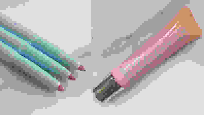 On the left: Three lip liners in different shades lay next to each other on a marble tabletop. On the right: A pink tube of lip gloss lays on a marble countertop.