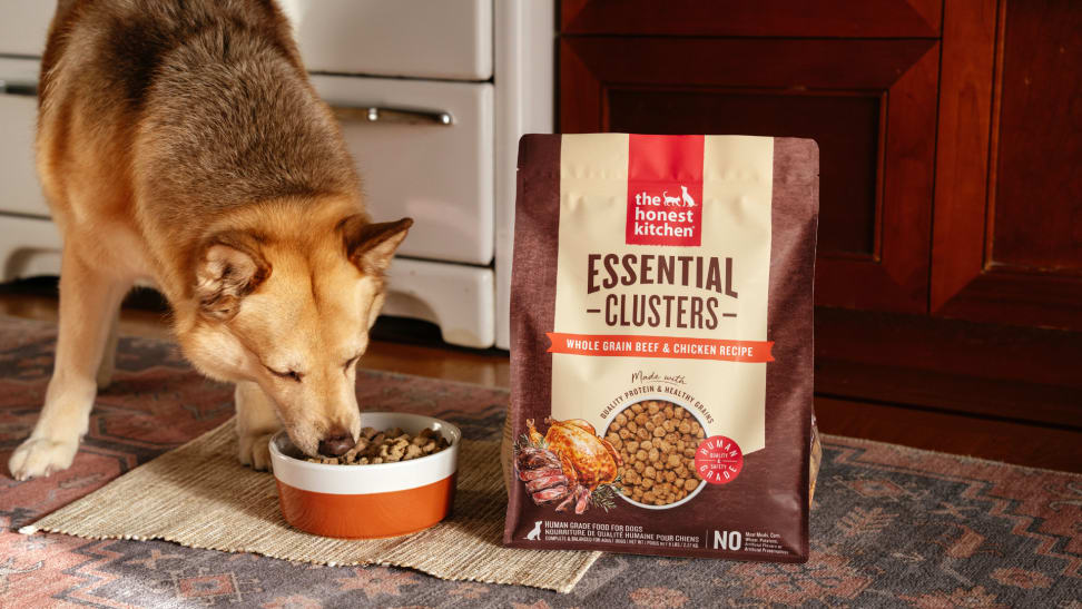 A dog eating from a bowl beside a bag of The Honest Kitchen dog food Essential Clusters whole grain beef and chicken recipe.