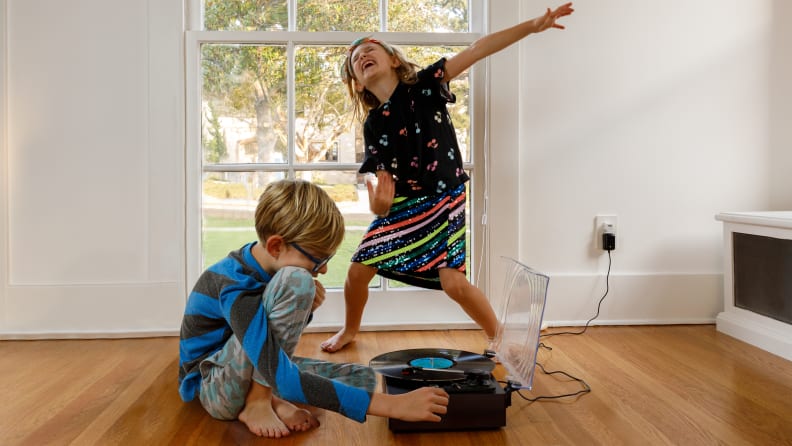 A girl wearing a sparkly skirt and black T-shirt sings along while a boy wearing blue pants plays a record on a record player.