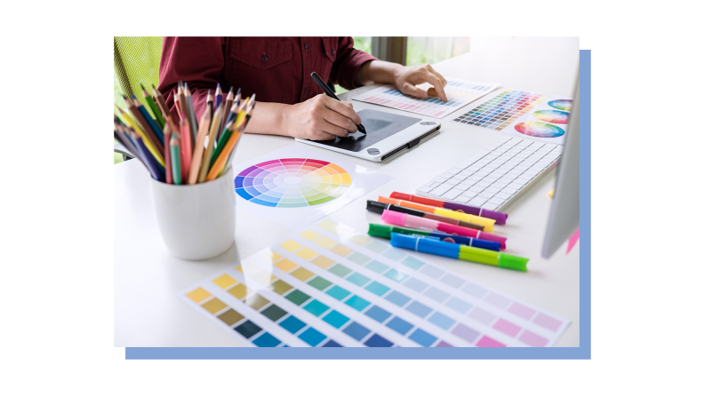 A freelance digital artist holds their stylus to a digital drawing board at a desk covered in color wheels, color pencils and markers.