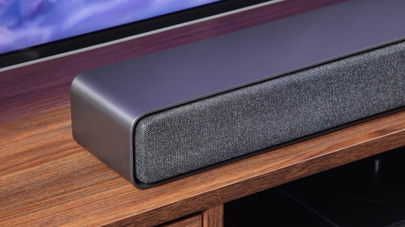 Close-up shot of the corner of the TCL Q Series Q6310 3.1-Channel Soundbar that has gray mesh material on front of speaker and a black exterior while it sits on top of wooden credenza..