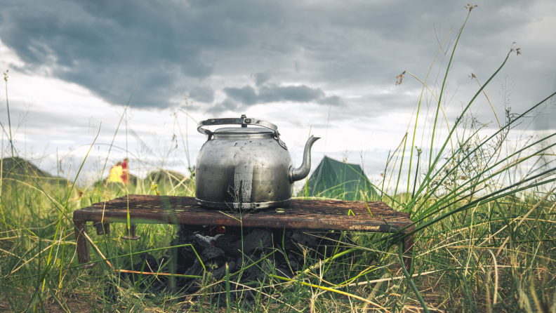 A metal kettle on top of a makeshift stove is seen in the middle of a grassland. A green tent is some distance behind.