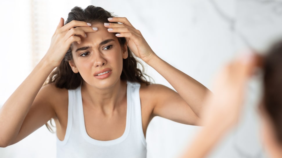 A person looking into a mirror and making an unhappy face and pinching at the skin on their forehead.