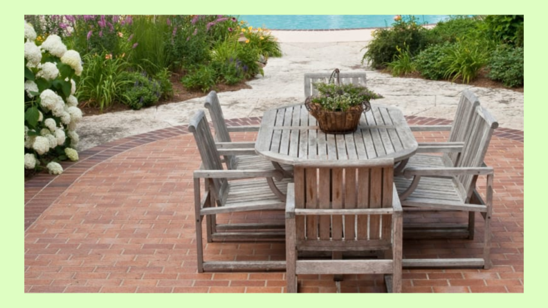 Wood patio set with plant sitting on brick patio flooring, surrounded by shrubs and flowers