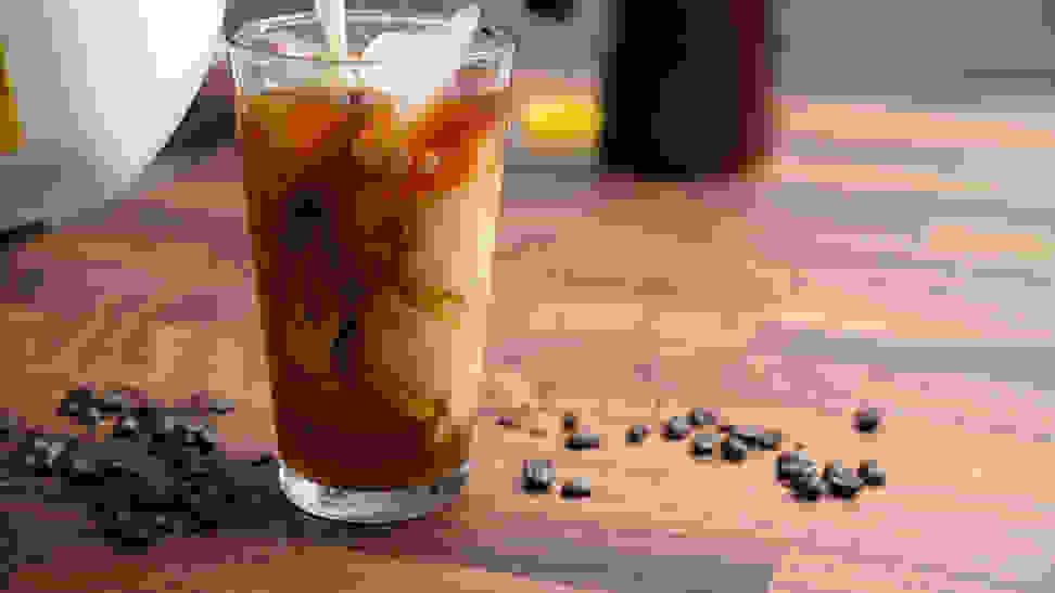 Milk pouring into a glass of cold brew coffee  surrounded by coffee beans sitting on a wood counter.