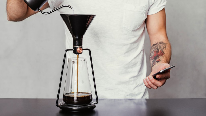 A person is making pour-over coffee using the Gina coffee maker while checking the Gina mobile app.