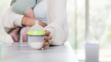 A parent reaches for a baby bottle filled with baby formula or breast milk while she holds her infant.