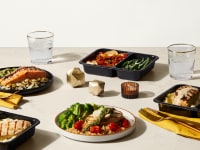 Multiple meals with seared chicken, salmon, vegetables on a beige table with geometric gold objects interspersed