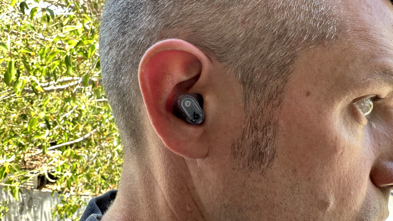 A man wearing a transparent Beats Studio Buds Plus earbud with trees in the background.