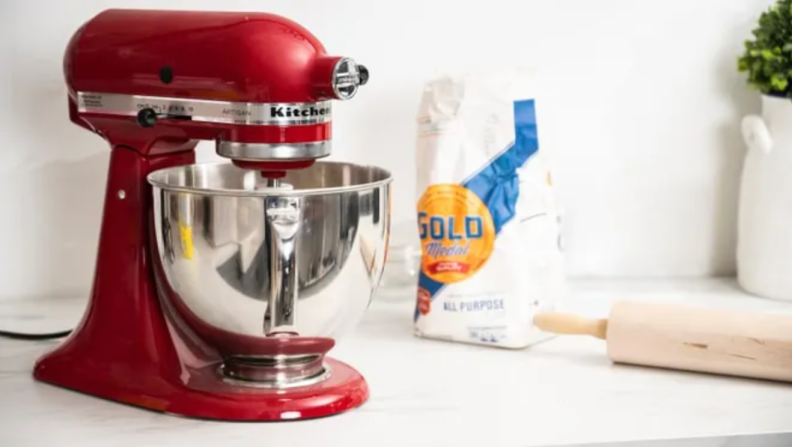 A red KitchenAid stand mixer on a kitchen countertop, next to a bag of flour and rolling pin.
