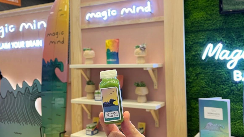 Person holding green wellness shot in front of Expo West display