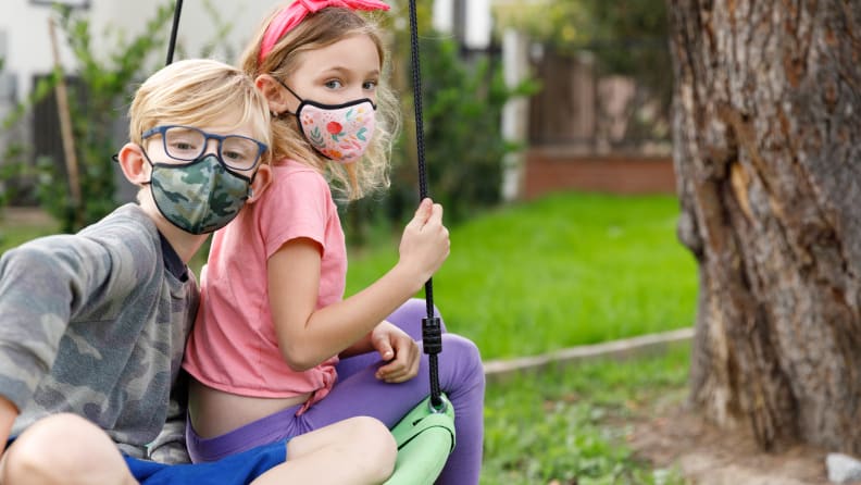 Two children sitting on a swing outside wearing face masks.