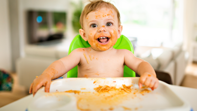 Baby with a face covered in pasta sauce sitting in a high chair.