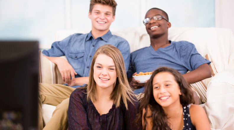A group of four teenagers sits in a living room and watches TV