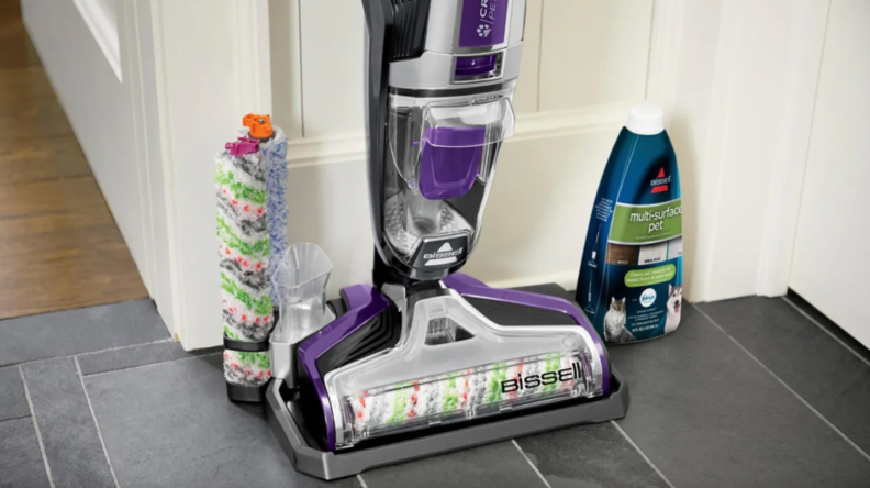 Bissell vacuum leaning against wall inside of home next to attachment pieces and cleaning solution.
