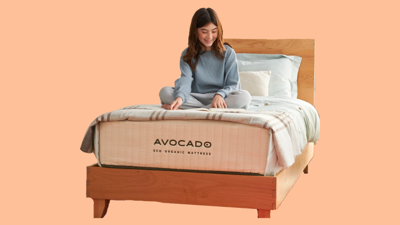 A woman sitting on top of a made bed showing the Avocado logo on the mattress, set against a pink background.