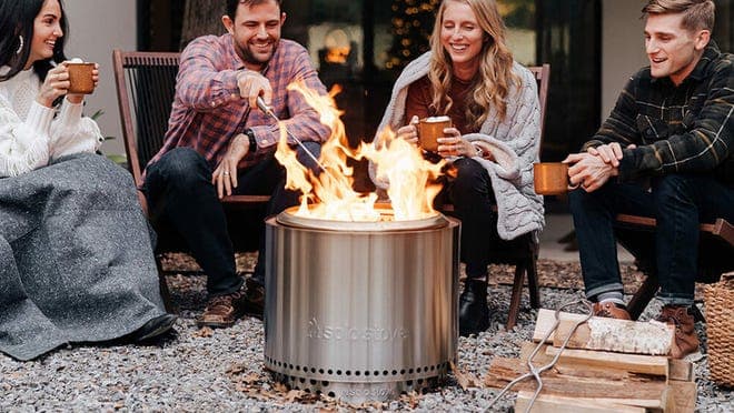 Four friends smiling while sitting around fire pit while roasting food on sticks and drinking out of mugs.