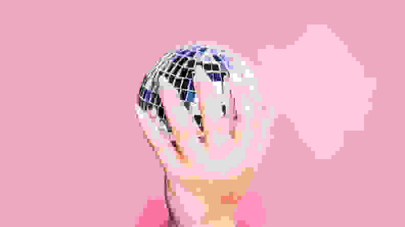 A hand wearing press-on nails holding a disco ball in front of a pink background.