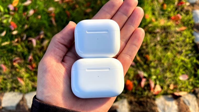 The all-white AirPods (3rd generation) case sits above the AirPods Pro case in a hand, in front of fall leaves and lush green grass.