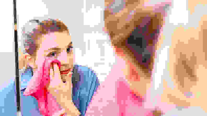 A woman stares into a large mirror and wipes her skin with a hot pink Makeup Eraser towel.