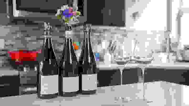 Three bottles of wine sit next to two champagne glasses on a marble countertop in a kitchen.