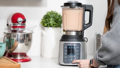A person blends a smoothie in a blender on a countertop surrounded by other kitchen items.