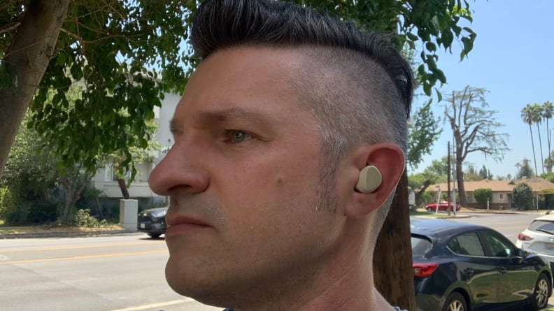 A man stands outside on a sidewalk looking off camera while wearing the gold beige Jabra Elite 5 earbuds.