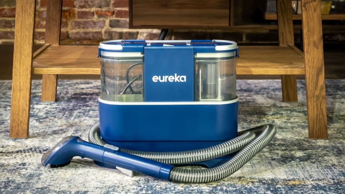 The Eureka NEY100 with hose attached on a patterned carpet in front of wooden table.
