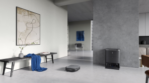 A square, black robot vacuum sits on a gray floor in the middle of a gray room. There is an abstract painting on a wall and a black bench with a blue blanket.