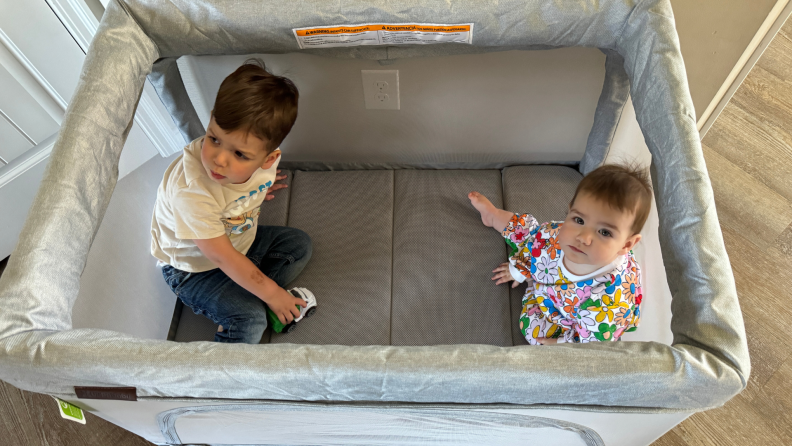 Two small toddlers sit on top of a small mattress inside of the Uppababy Remi while looking up at the camera.