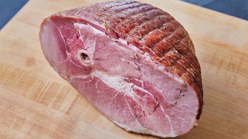 A bone-in honey baked ham sits on a light wood cutting board.