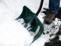 A close-up of a snow shovel digging into a snow bank. The person holding it is attempting to clear a walkway that's covered in a few inches of snow. .