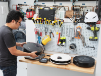 Person standing at work bench with assorted tools hanging along pegboard wall while inspecting the weight and dimensions of several small robot vacuums inside of testing lab with stacked washer and dryer combo.